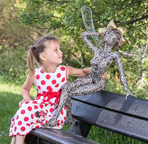Image shows a girl sitting on a bench to admire one of the wire fairy sculptures at The Trentham Estate